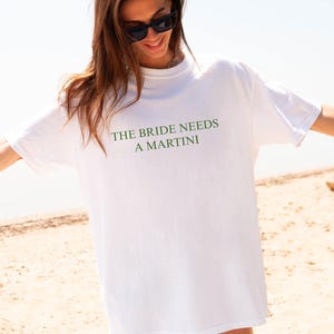 May include: White t-shirt with the text "THE BRIDE NEEDS A MARTINI" in green. The shirt is worn by a person on a beach, with sunglasses and arms outstretched. A camera and beach bag are visible.