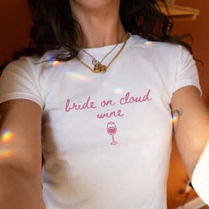 May include: White t-shirt with the pink text "Bride on cloud wine" and a wine glass graphic. The shirt is a crew neck style.