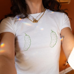 May include: White t-shirt with two green pickle illustrations. The shirt is short-sleeved and appears to be made of cotton. The pickles are drawn with a simple outline and are positioned on the front of the shirt.