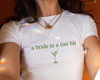Może przedstawiać: Biała koszulka z napisem "a bride in a tini bit" i zieloną ilustracją kieliszka do martini.