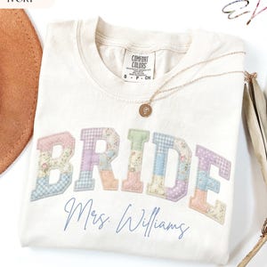 May include: An ivory t-shirt featuring the word BRIDE in large, patchwork-style letters with floral and gingham patterns in pastel colours. Below the word, the text Mrs. Williams is printed in a blue script font. A gold necklace rests on the shirt.