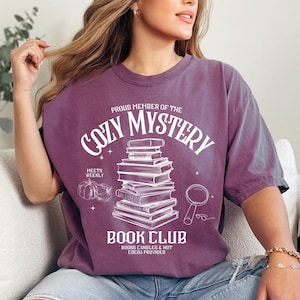 Peut inclure: Un t-shirt violet avec un motif graphique blanc qui dit "Proud Member of the Cozy Mystery Book Club" avec une pile de livres, une loupe et des citrouilles. Le texte indique également "Meets Weekly" et "Books, Candles & Hot Cocoa Provided."
