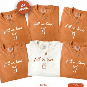 May include: Five short-sleeved crew neck t-shirts. Four shirts are a muted orange color with white text reading fall in love and a white bow graphic. One shirt is ivory with red text reading fall in love and a red engagement ring graphic.