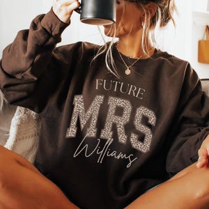 May include: A brown sweatshirt with the text "FUTURE MRS Williams" in white and leopard print. A black mug is held up. The sweatshirt is worn by a person sitting on a couch.
