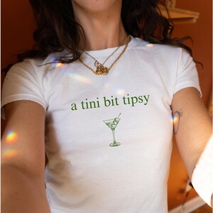 May include: White t-shirt with the text "a tini bit tipsy" and a green illustration of a martini glass with olives.