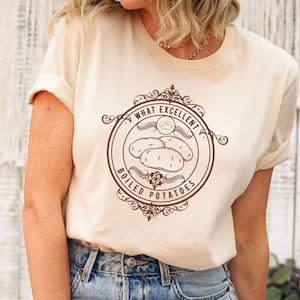 May include: A woman wearing a light beige t-shirt with a brown graphic of a vintage-style logo that says "What Excellent Boiled Potatoes". The graphic features a drawing of three potatoes. The woman is wearing light blue jeans with distressed details.
