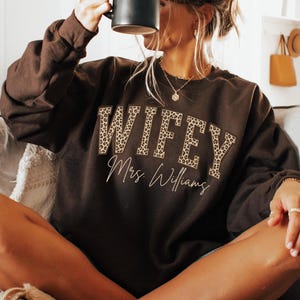 May include: A brown sweatshirt featuring the word "WIFEY" in leopard print and the name "Mrs. Williams" in cursive script. A black mug is held up. The sweatshirt is a relaxed fit.