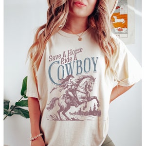 K&ouml;nnte beinhalten: Cremefarbenes T-Shirt mit einer Grafik einer Cowgirl auf einem Pferd und dem Text "Save A Horse Ride A Cowboy" in Blau und Braun. Das Design beinhaltet eine Stadtsilhouette. Das Shirt hat eine lockere Passform.