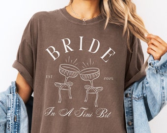 May include: A brown t-shirt with the text "BRIDE EST 2025" and a graphic of two champagne flutes with bows. The text "In A Tini Bit" is below the graphic.