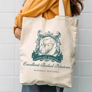 May include: Cream-colored tote bag with a teal and white graphic of two potatoes inside an ornate frame. The text reads "Exemplary Vegetable" and "Excellent Boiled Potatoes". The bag has a simple design with long handles.