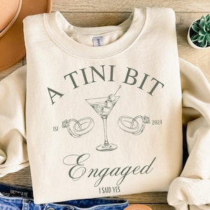 May include: A beige sweatshirt with the text "A TINI BIT Engaged I SAID YES" and a martini glass with olives and two rings. The year 2024 and the word "EST" are also on the sweatshirt.