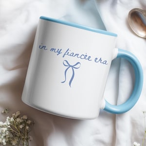 May include: White ceramic mug with a light blue rim and handle. The mug features the text "in my fiancée era" in light blue script, along with a bow graphic. The mug is on a white surface.