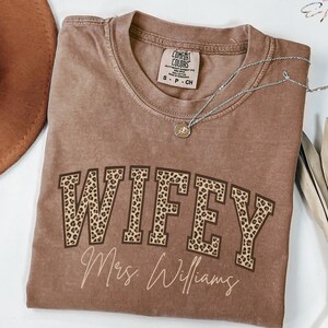 May include: A brown Comfort Colors t-shirt with the word "WIFEY" in large, leopard print letters. Below the word "WIFEY" is the name "Mrs. Williams" in a cursive font. A silver necklace with a small pendant is on the shirt.