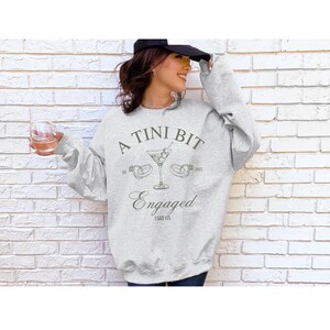 May include: A gray sweatshirt with the text "A Tini Bit Engaged I Said Yes" and a martini glass with olives and two rings on the stem.
