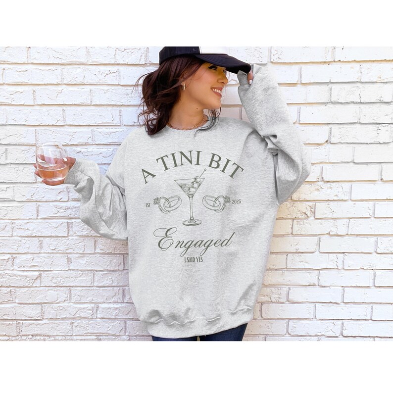 May include: A gray sweatshirt with the text "A Tini Bit Engaged I Said Yes" and a martini glass with olives and two rings on the stem.