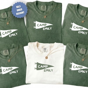 May include: Several green and white t-shirts, each displaying the text "CAMP EMILY" in a pennant design. The shirts are accessorized with gold-coloured necklaces. A blue circle with "BULK ORDER DISCOUNT" is visible in the top left.
