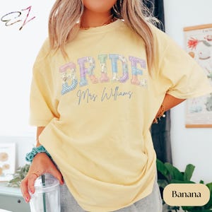 May include: A pale yellow short-sleeve t-shirt featuring the word BRIDE in large, blocky, multi-coloured pastel gingham and floral patterned letters, with the text Mrs. Williams written in a dark blue cursive script underneath.