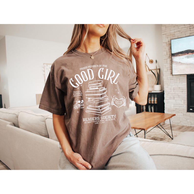 May include: A brown t-shirt with a white graphic design featuring a stack of books, a skull, and the text "Proud Member of the Good Girl Readers Society We Don't Watch Pork We Read It Like A Lady".
