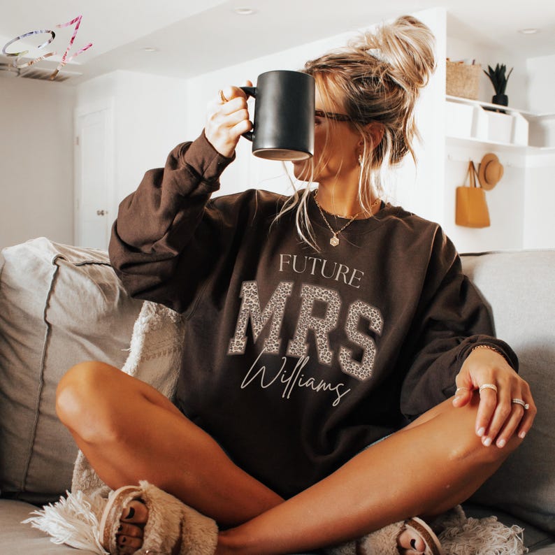 May include: A brown sweatshirt with the text "FUTURE MRS Williams" in white and leopard print. A black mug is held up. The sweatshirt is worn by a person sitting on a couch.