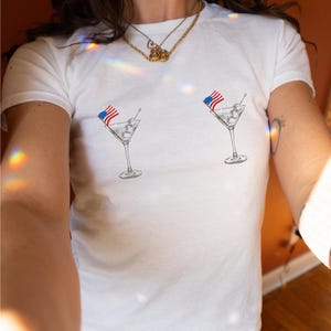 May include: White t-shirt with a graphic of two martini glasses with American flags on the stems.