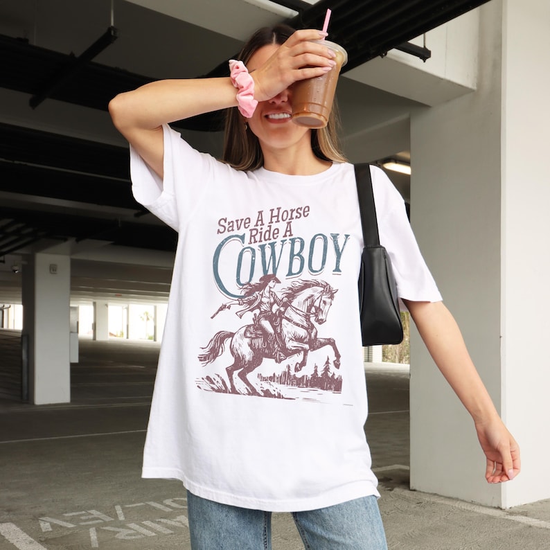 K&ouml;nnte beinhalten: Wei&szlig;es T-Shirt mit einem Aufdruck "Save A Horse Ride A Cowboy". Das Design zeigt einen Cowboy auf einem Pferd in Sepiat&ouml;nen. Das Shirt hat eine lockere, &uuml;bergro&szlig;e Passform.