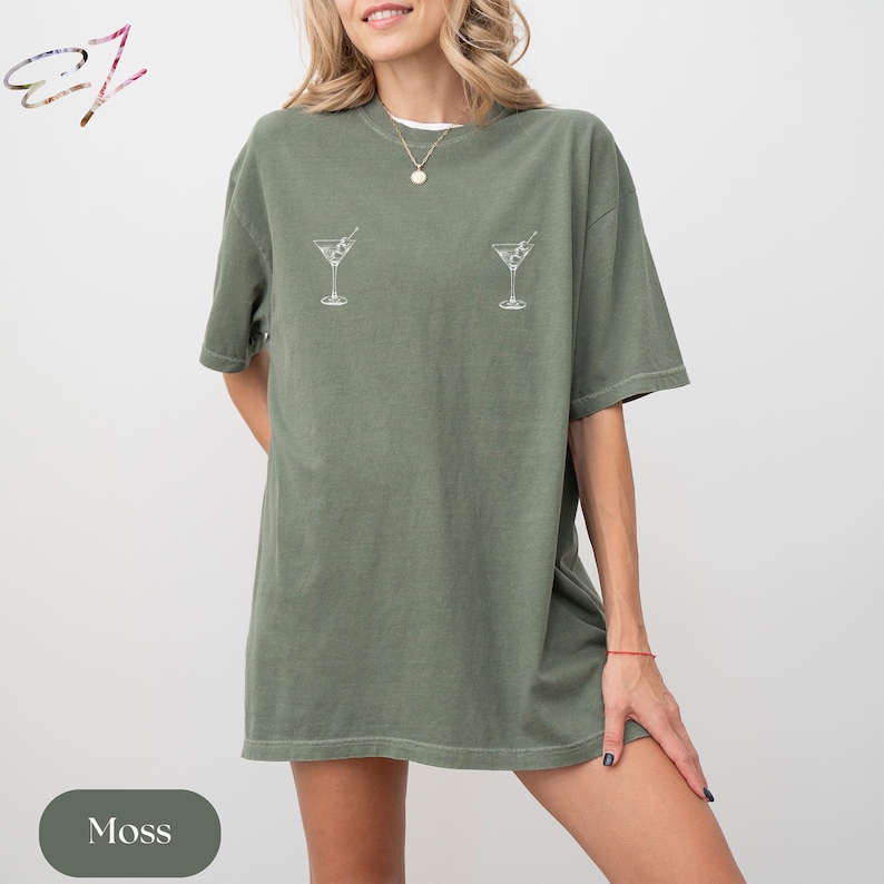 May include: An oversized moss green t-shirt featuring two small, white line-art illustrations of martini glasses positioned on the upper chest area. The garment has short sleeves, a crew neckline, and a relaxed, loose-fitting silhouette.