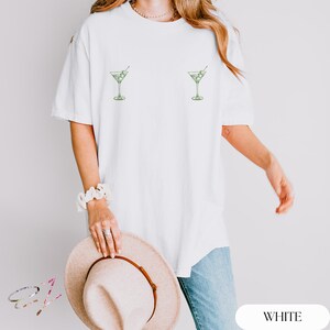 May include: A white short-sleeved t-shirt featuring two small, green line-art illustrations of martini glasses with olives, positioned symmetrically on the chest area. The shirt has a crew neckline and a relaxed, loose-fitting silhouette.