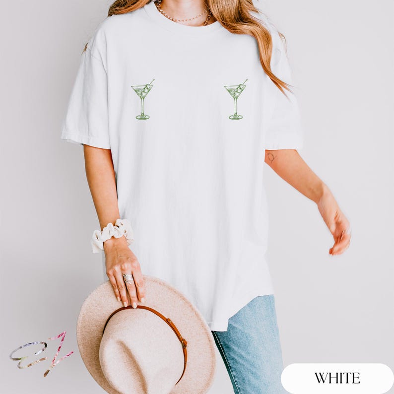 May include: A white short-sleeved t-shirt featuring two small, green line-art illustrations of martini glasses with olives, positioned symmetrically on the chest area. The shirt has a crew neckline and a relaxed, loose-fitting silhouette.