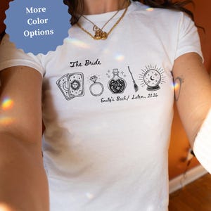 May include: A white short-sleeve t-shirt featuring a black graphic print of tarot cards, a diamond ring, a potion bottle, a broom, and a crystal ball. The text reads The Bride and Emily's Bach/ Salem, 2026 in a cursive and print font style.