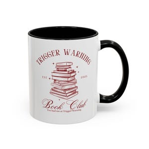 May include: White ceramic mug with a black interior and handle. The mug features the text "TRIGGER WARNING Book Club" in burgundy, along with a stack of books graphic. The text "You had me at Trigger Warning" is also included.