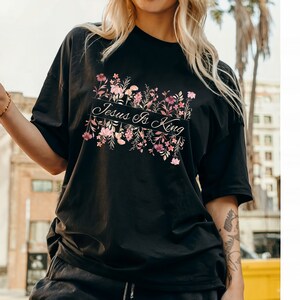 Pode incluir: Camiseta preta com a frase "Jesus Is King" em um design floral. O design apresenta flores rosa e roxas. A camiseta tem mangas curtas e um corte folgado.