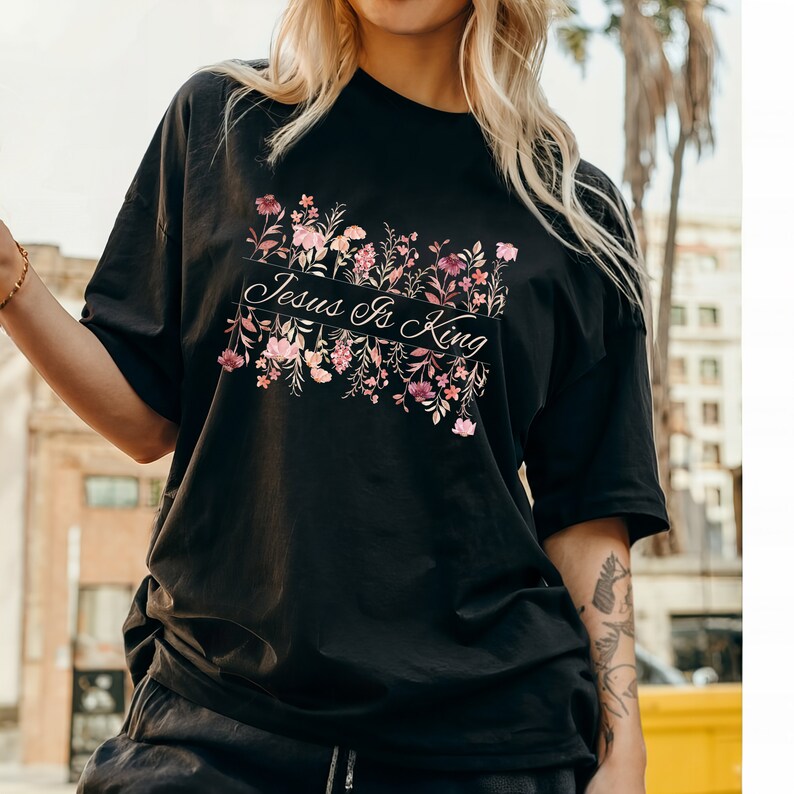 Pode incluir: Camiseta preta com a frase "Jesus Is King" em um design floral. O design apresenta flores rosa e roxas. A camiseta tem mangas curtas e um corte folgado.