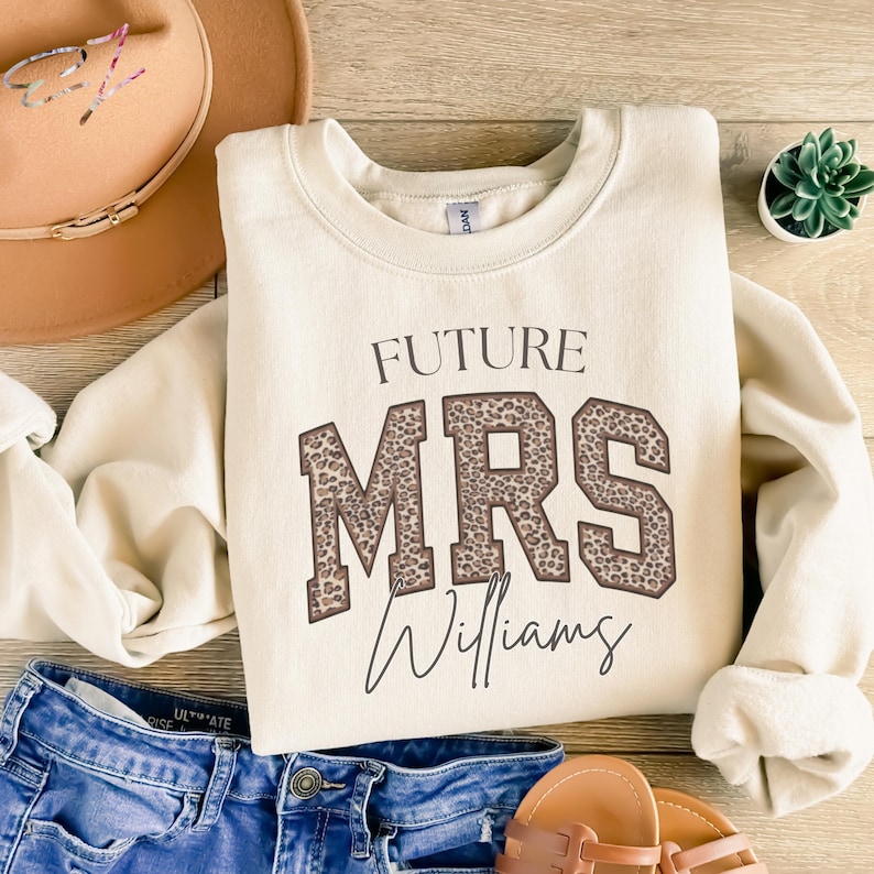 May include: Cream-colored sweatshirt with the text "FUTURE MRS Williams" in a combination of leopard print and script font. The sweatshirt is paired with blue denim shorts, a brown hat, and sandals.
