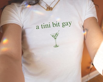 Op de afbeelding: Wit T-shirt met de groene tekst "a tini bit gay" en een groene martini glas illustratie. Het shirt heeft korte mouwen en een ronde hals.