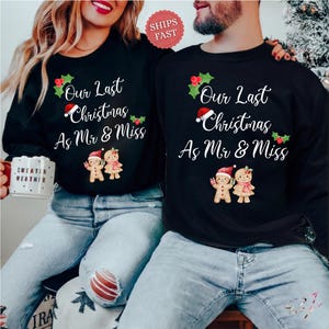 Op de afbeelding: Twee zwarte sweaters met ronde hals en de witte tekst Our Last Christmas As Mr & Miss, versierd met rode en groene hulst, een kerstmuts en twee peperkoekmannetjes. E&eacute;n sweater wordt vastgehouden met een witte mok met de tekst Sweater Weather.