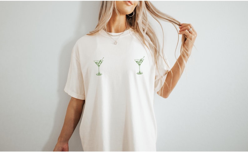 May include: An off-white short-sleeve t-shirt featuring two small, green martini glass graphics printed on the chest area. Each glass contains liquid and olives on a toothpick. The shirt has a crew neckline and a relaxed, loose-fitting silhouette.