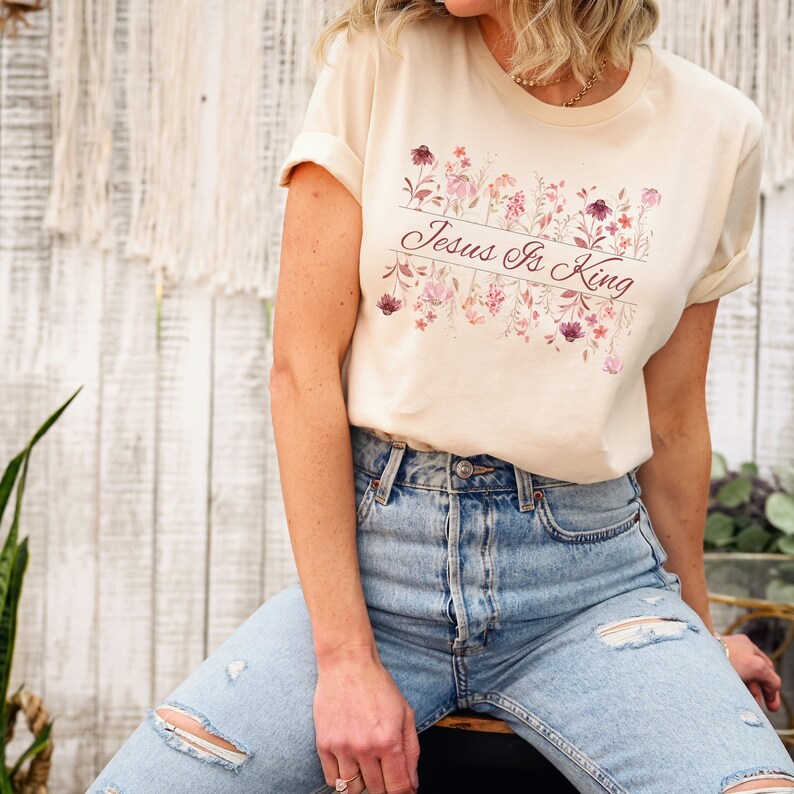 Pode incluir: Camiseta creme com mangas enroladas. A camisa apresenta um design floral em tons de rosa e roxo, com a frase "Jesus Is King" impressa em letra cursiva. A modelo est&aacute; usando jeans azul claro.