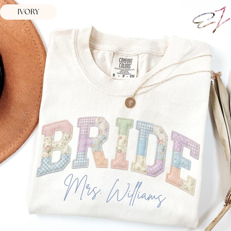 May include: An ivory t-shirt featuring the word BRIDE in large, patchwork-style letters with floral and gingham patterns in pastel colours. Below the word, the text Mrs. Williams is printed in a blue script font. A gold necklace rests on the shirt.