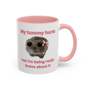 May include: White ceramic mug with a pink rim and handle. The mug features a cartoon image of a sad cat with a pink bow and the text "My tummy hurts but I'm being really brave about it".