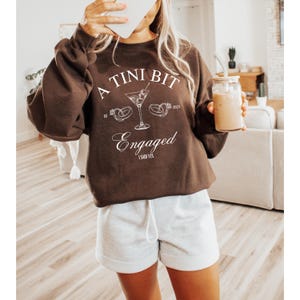 May include: A brown sweatshirt with the text "A Tini Bit Engaged" and a martini glass with two rings on the rim. The text "Est 2021" is also on the sweatshirt.