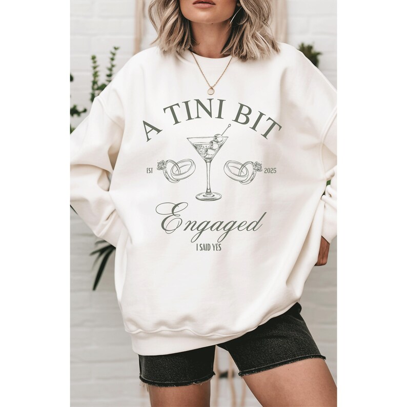 May include: A white sweatshirt with the text "A TINI BIT ENGAGED I SAID YES" and a martini glass with olives and two rings on either side of the glass. The text "EST 2025" is below the rings.