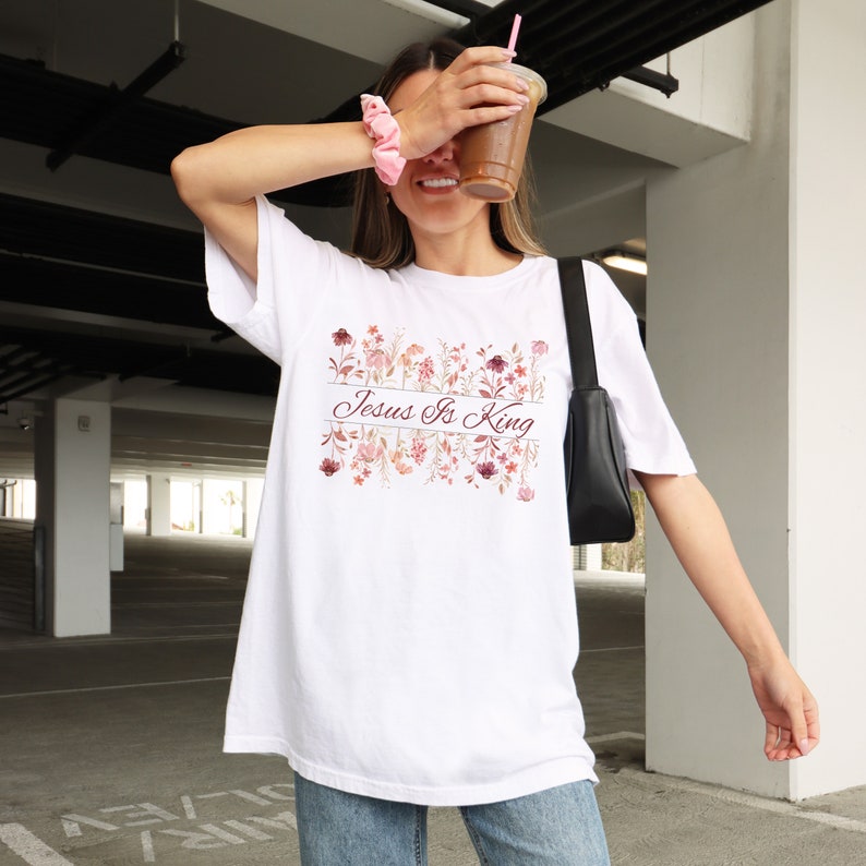 Pode incluir: Camiseta branca com a frase "Jesus Is King" em um design floral. O design apresenta flores em tons de rosa, vermelho e p&ecirc;ssego. A camisa &eacute; de um estilo casual e confort&aacute;vel.