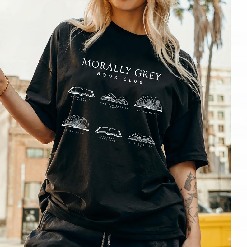 May include: Black t-shirt featuring the white text "MORALLY GREY BOOK CLUB". Underneath are six open book illustrations with titles: "ENEMIES TO LOVERS", "WHO DID THIS TO YOU", "FATED MATES", "SLOW BURN", "TRIGGER WARNINGS", and "I'LL KILL FOR YOU".