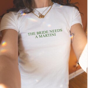 May include: White t-shirt with the green text "THE BRIDE NEEDS A MARTINI". The shirt is a crew neck style and is made of a soft material. The shirt is paired with blue jeans.