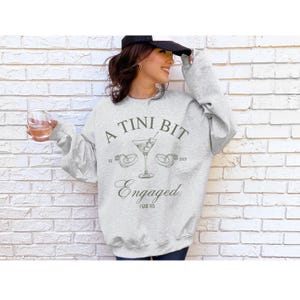 May include: A gray sweatshirt with the text "A Tini Bit Engaged" and a martini glass with olives and two rings. The text also includes "Est 2021" and "2023" and "I Said Yes".
