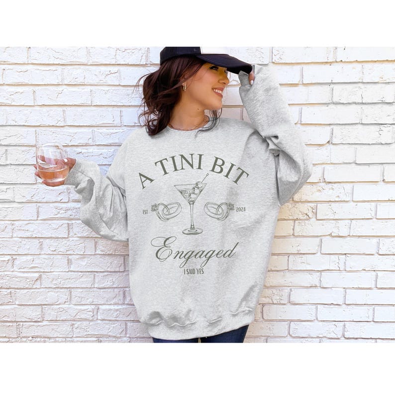 May include: A gray sweatshirt with the text "A Tini Bit Engaged" and a martini glass with olives and two rings. The text also includes "Est 2021" and "2023" and "I Said Yes".