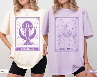May include: Two oversized t-shirts, one ivory and one orchid, each featuring a tarot card design in purple. The ivory shirt displays "THE BRIDE" with a woman in a gown. The orchid shirt shows "THE COVEN" with hands and crystals.