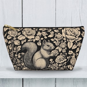 May include: Black cosmetic bag with a floral and squirrel design. The bag features a detailed illustration of a squirrel holding a rose, surrounded by roses and foliage in cream and black. It has a gold zipper.