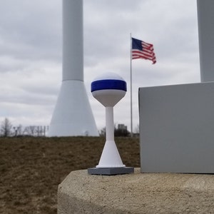May include: A white water tower with a blue band and the text "GAS CITY" and "gascityindian.com". A miniature white water tower with a blue band is in the foreground. The miniature water tower is sitting on a gray concrete surface.