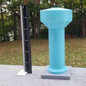 Puede incluir: Un modelo de torre de agua de plástico azul claro con una base cilíndrica y una parte superior redondeada. El modelo mide aproximadamente 15 cm de alto y se encuentra sobre una base gris.
